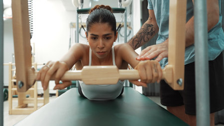 Instructor assisting a client in a bar-based exercise on a Pilates Cadillac table. Woman performing swan diving on the cadillac for improving upper body strength and core engagement at gym. Habituate.の写真素材