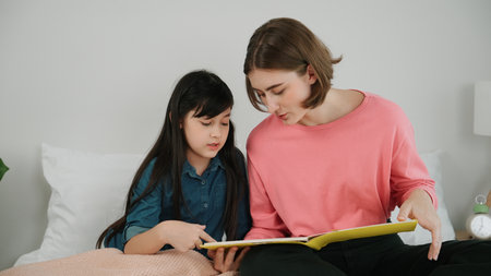 Caucasian mother reading story book to cute daughter listening story. Asian girl pointing while...