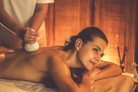 Hot herbal ball spa massage body treatment, masseur gently compresses herb bag on woman body. Tranquil and serenity of aromatherapy recreation in warm lighting of candles at spa salon. Quiescentの写真素材