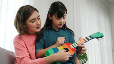 Happy girl playing ukulele while caucasian mom teaching and explain about acoustic music at home. Cute child learning about instrument. Happy caucasian mother and girl spend time together. Pedagogy.の写真素材