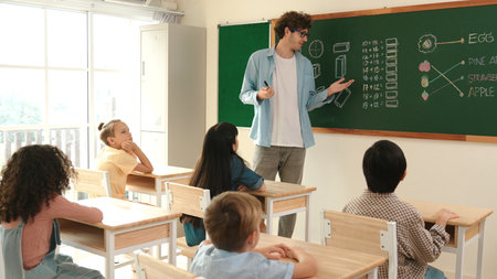 Diverse elementary student walking in classroom while teacher looking at child. Cute children attend classroom while professional instructor prepare for teaching and standing at blackboard. Pedagogy.の写真素材