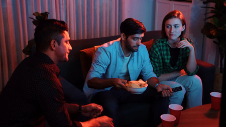 Caucasian father and mother talking with colleague while pick up snack bowl to friend. Happy family sitting at seat in living room while enjoy watching movie with caucasian man at home.の写真素材