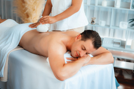 Hot herbal ball spa massage body treatment, masseur gently compresses herb bag on man body. Tranquil and serenity of aromatherapy recreation in day lighting ambient at spa salon. Quiescentの写真素材