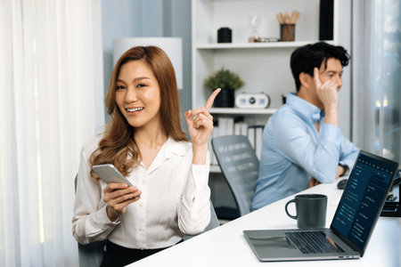Profile smiling beautiful Asian businesswoman looking camera, using phone for advertisement at modern office on working desk casual day. Blurry background man colleague analyzing data on pc. Infobahn.の写真素材