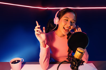Host channel of young beautiful Asian broadcaster listening the music on social media with listeners, wearing pastel headphones, using microphone for singing at neon light indoor studio. Stratagem.の写真素材