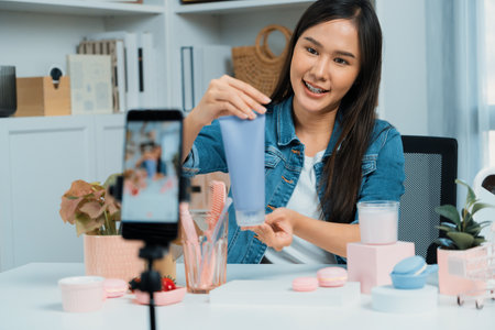 Young beautiful Asian girl reviewing moisturizing body lotion cream product to make glow skin testing on arm on social media online live recording smartphone with bestseller at modern room.の写真素材
