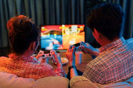 Couple gamer with joysticks playing fighting video game together on tv screen, getting challenge level at back side view, sitting sofa at neon light color living room at modern comfy home. Infobahn.の写真素材