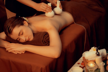 Hot herbal ball spa massage body treatment, masseur gently compresses herb bag on woman body. Tranquil and serenity of aromatherapy recreation in warm lighting of candles at spa salon. Quiescentの写真素材