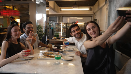 Woman taking sefie using smartphone with friends posing with special cocktail drinks surrounded by grill and fried meat special menu featuring unique chef table menu on table at restaurant. Vinosity.の写真素材