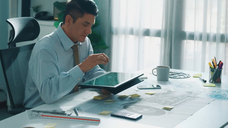 Professional engineer looking at phone while working on tablet with green screen at meeting table...