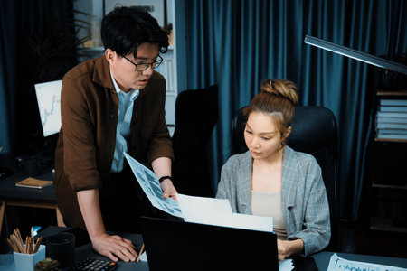 Serious face working on laptop of businesswoman with coworker giving overwork paperwork at over late...