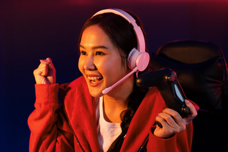 Host channel of smiling beautiful Asian girl streamer with joystick playing with raising fist up winning Esport skilled team players wearing headphones pastel media online in neon room. Stratagem.の写真素材