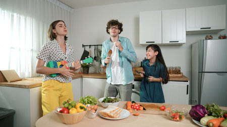 Caucasian skilled father, mother and asian daughter making breakfast while dancing together. Skilled mom playing ukulele while preparing vegetable at modern kitchen. Healthy food concept. Pedagogy.の写真素材