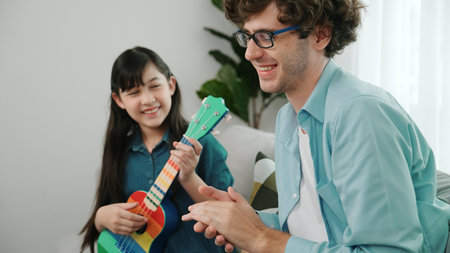 Back view of happy father looking at smart daughter playing ukulele and singing song. Happy dad motivated and encouraged school girl enjoy playing guitar. Family recreation. Pedagogy.の写真素材