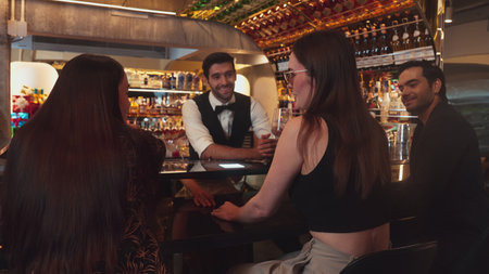 Smart bartender with VIP customer at luxurious bar greeting new beautiful guest at counter in engaging lively conversation at back view in lighting atmosphere at warm nightclub.の写真素材