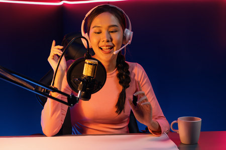 Smiling creative young beautiful Asian influencer talking in channel with positive podcast speech at...