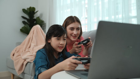 Happy girl playing game and sitting while mom lies on sofa by using laptop screen. Parent and girl...
