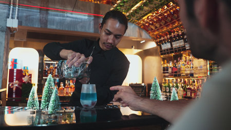 Bartender pouring mixed blue lagoon cocktail liqueur to cooling glass while guest talking friends waiting drink, decorated pine light christmas theme making special festive tasty in couple. Vinosity.の写真素材