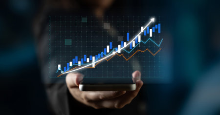 A businessman holds a smartphone displaying a digital graph with upward trends. This image captures...