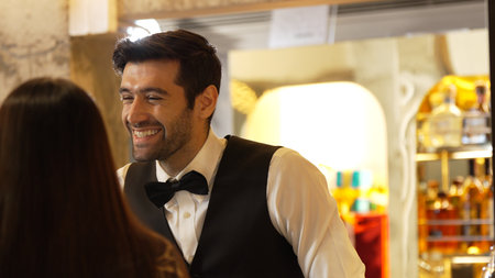 Smiling smart bartender providing excellent service to beautiful guest woman at back side vibrant nightlife party atmosphere in engaging lively conversation with VIP customer at nightclub.の写真素材