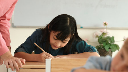 Asian girl writing or taking a note while teacher checking classwork. Caucasian teacher help happy...