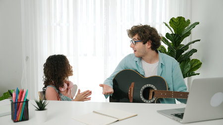 Skilled caucasian father playing guitar to relax daughter after learning engineering. Cute schoolgirl with headphone sitting at table with laptop while listen acoustic music. Pedagogy.の写真素材