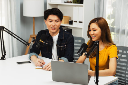 Smiling radio influencer hosts with channel social media online live streaming with talking topic collective question on script note and laptop to listeners at morning time at studio record.の写真素材