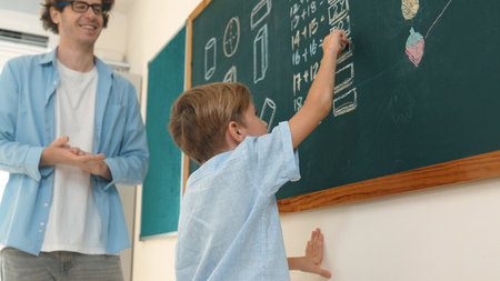 Happy caucasian boy writing answer at blackboard and thinking about theory while standing in front...