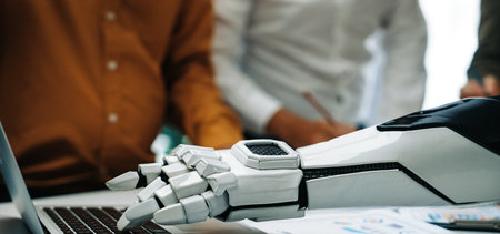 A robotic hand interacts with a laptop in a professional office setting, showcasing the blend of technology and human collaboration in modern workplaces.の写真素材