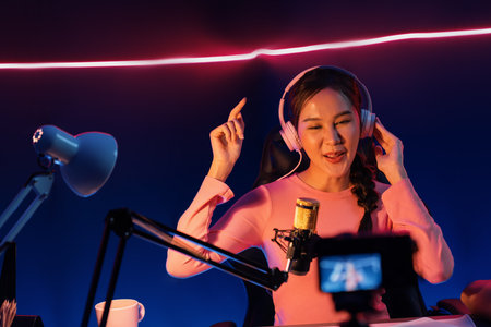 Selective focus camera screen in host channel of beautiful Asian listening the music on social media with listener, wearing pastel headphones on blurred background using mic at neon studio.の写真素材