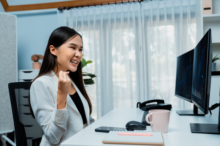 Successful young Asian businesswoman earning highest profit with fist up in exchange stock market on dynamic business graph on pc screens at modern office, analyzing financial technology. Stratagem.の写真素材