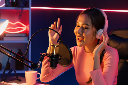 Host channel of young beautiful Asian broadcaster listening the music on social media with listeners, wearing pastel headphones, using microphone for singing at neon light indoor studio. Stratagem.の写真素材