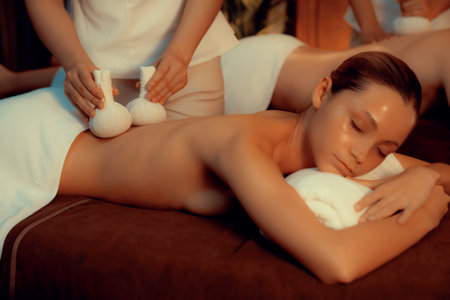 Hot herbal ball spa massage body treatment, masseur gently compresses herb bag on couple customer body. Serenity of aromatherapy recreation in warm lighting of candles at spa salon. Quiescentの写真素材
