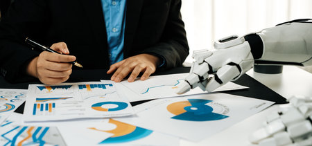 A business professional assesses financial data while a robotic hand extends towards documents, highlighting the merge of technology and human expertise in analysis.の写真素材