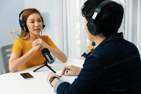 Smiling radio influencer host wearing headphone interviewing woman speaker sharing information to listeners on social media online website live streaming channel at morning at studio record.の写真素材