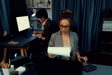 Businesswoman with stressful face looking document paperwork file while coworker trading stock market at back at over late night time at modern office, working on startup company project. Infobahn.の写真素材
