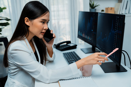 Young Asian businesswoman calling to broker, pointing with pen in increased exchange stock market on pc to invest high profit at modern office, investing high profit in wealth.の写真素材