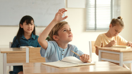 Caucasian smart boy raising hand for asking and answering teacher surrounded by diverse children studying at classroom while teacher walking pass at elementary classroom. Education concept. Pedagogy.の写真素材