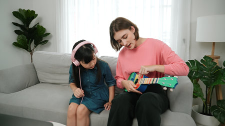 Asian cute girl listening to music from laptop by using headphone while lovely mom playing ukulele while teaching young daughter playing acoustic music. Cute child learning about instrument. Pedagogy.の写真素材