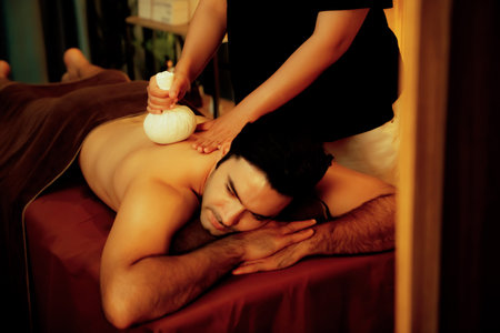 Hot herbal ball spa massage body treatment, masseur gently compresses herb bag on man body. Tranquil and serenity of aromatherapy recreation in warm lighting of candles at spa salon. Quiescentの写真素材
