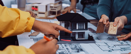 A diverse group of professionals collaborates on an architectural design project using a house model, colorful materials, and blueprints in a bright office setting.の写真素材