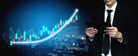 A business professional examines financial data on a tablet while displaying a growing graph against...