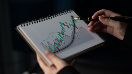 Hands showcase an open notepad displaying a stock market chart, while a pen highlights details on the candlestick graph, emphasizing financial analysis in an office setting. Imputeの写真素材