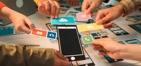 A vibrant scene of hands collaborating in a creative session around a mobile application design. Various colorful icons surround a smartphone mockup, illustrating teamwork and innovation.の写真素材