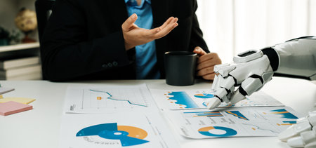 A human and a robot interact at an office desk, examining charts and data reports together while...