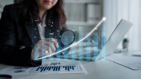 A focused businesswoman reviews financial data with growth charts on her laptop, representing...