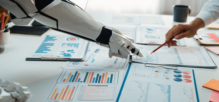 This image captures a robotic arm working alongside a human hand over various business charts and...