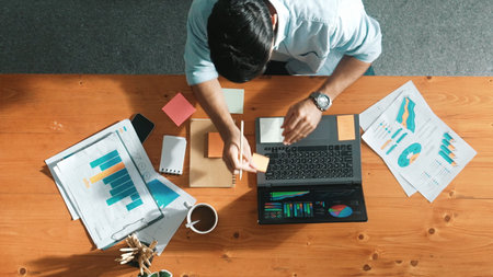 Skilled businessman writing idea on colorful sticky notes while looking at data analysis. Caucasian project manager working on laptop while taking a note in sticky notes at meeting room.の写真素材