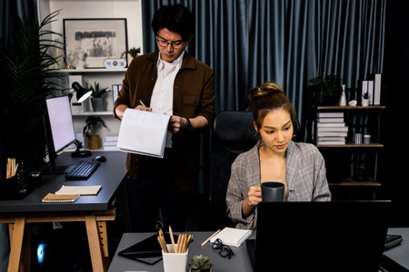 Businesswoman looking on laptop for thinking marketing online project while drinking coffee with coworker working at back side holding paperwork at late night time at modern office.の写真素材