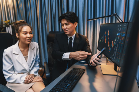 Asian businessman showing stock market application on mobile phone to partner pointing by pen,...